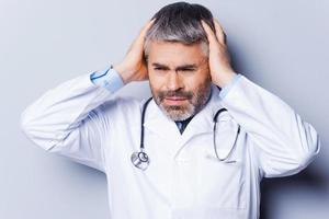 stressato medico. frustrato maturo medico Tenere mani nel capelli e guardare lontano mentre in piedi contro grigio sfondo foto