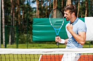 godendo vittoria. lato Visualizza di contento giovane uomo nel polo camicia Tenere tennis racchetta e Esprimere a gesti mentre in piedi su tennis Tribunale foto