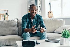 moderno giocatore. bello giovane africano uomo giocando video gioco e sorridente mentre seduta in casa foto