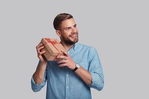 grande regalo. bello giovane uomo tremante regalo scatola e sorridente mentre in piedi contro grigio sfondo foto