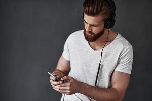 ambientazione su il suo elenco di riproduzione. bello giovane uomo ascoltando musica e utilizzando il suo inteligente Telefono mentre in piedi contro grigio sfondo foto