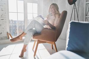 godendo lettura. bellissimo giovane donna lettura un' rivista mentre seduta nel il poltrona a casa foto