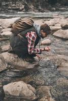 rinfrescante con freddo acqua. bello giovane moderno uomo potabile acqua a partire dal il fiume mentre escursioni a piedi nel montagne foto