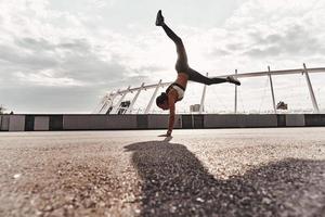 energia e equilibrio. pieno lunghezza di moderno giovane donna nel gli sport capi di abbigliamento fare handstand mentre esercizio all'aperto foto