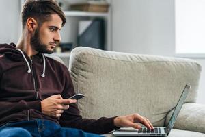 multitasking. lato Visualizza di concentrato giovane uomo utilizzando il suo il computer portatile e Tenere inteligente Telefono mentre seduta su divano a casa foto