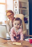 moderno Lavorando madre. poco bambino ragazza guardare a telecamera mentre strisciando su sua madri posto di opera foto