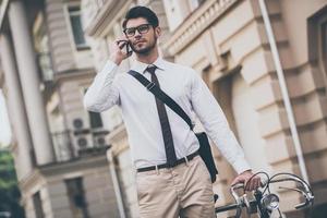 vedere voi nel ufficio fiducioso giovane uomo nel bicchieri parlando su mobile Telefono e Tenere mano su il suo bicicletta mentre in piedi all'aperto foto