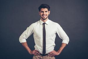 migliore Sorridi. fiducioso giovane bello uomo conservazione braccia akimbo e guardare a telecamera con Sorridi mentre in piedi contro grigio sfondo foto