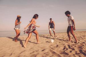 la spesa spensierato tempo insieme. gruppo di allegro giovane persone giocando con calcio palla su il spiaggia con mare nel il sfondo foto