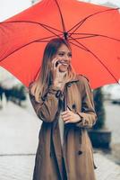 simpatico per sentire a partire dal voi attraente giovane sorridente donna trasporto ombrello e parlando su il mobile Telefono mentre in piedi su il strada foto