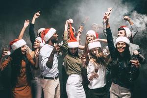 nuovo anno festa con gli amici. gruppo di bellissimo giovane persone nel Santa cappelli danza e guardare contento foto