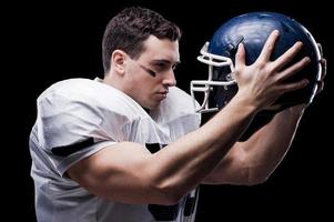 combattente spirito. americano calcio giocatore Tenere calcio casco nel davanti di il suo viso e guardare a esso mentre in piedi contro nero sfondo foto