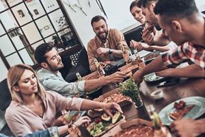 delizioso cena. gruppo di giovane persone nel casuale capi di abbigliamento mangiare e sorridente mentre avendo un' cena festa in casa foto