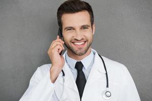 sempre pronto per Aiuto voi. gioioso giovane medico nel bianca uniforme sorridente e parlando su il mobile Telefono mentre in piedi contro grigio sfondo foto