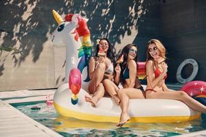 attraente giovane donne nel costumi da bagno sorridente e mangiare anguria mentre galleggiante su gonfiabile unicorno nel nuoto piscina all'aperto foto