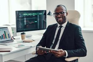 concentrato giovane africano uomo nel vestito formale utilizzando moderno tecnologie e sorridente mentre Lavorando nel il ufficio foto