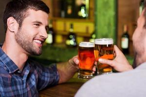 Venerdì notte fuori. Due allegro giovane uomini tostatura con birra e sorridente mentre seduta insieme a il bar contatore foto