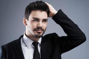 Usato per Guarda Perfetto. ritratto di fiducioso giovane uomo nel vestito formale Tenere mano nel capelli e guardare lontano mentre in piedi contro grigio sfondo foto