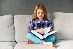 tempo per fiaba. bellissimo poco ragazza lettura libro mentre seduta su il divano nel loto posizione a casa foto