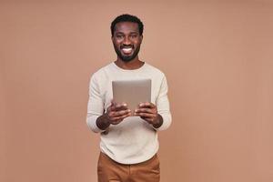 bello giovane africano uomo nel casuale capi di abbigliamento utilizzando digitale tavoletta e sorridente foto