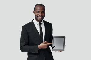 fiducioso giovane africano uomo nel vestito formale puntamento copia spazio su digitale tavoletta mentre in piedi contro grigio sfondo foto