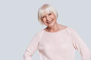 bellezza ha no età. bellissimo anziano donna guardare a telecamera e sorridente mentre in piedi contro grigio sfondo foto