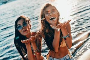 condivisione divertimento con voi. superiore Visualizza di Due attraente giovane donne nel costumi da bagno sorridente e soffiaggio coriandoli mentre in piedi vicino il fiume all'aperto foto