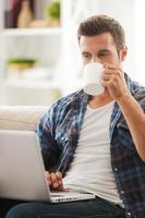 ottenere il ultimo notizia in linea. bello giovane uomo Lavorando su il computer portatile e potabile caffè mentre seduta su divano foto