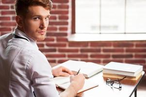 fiducioso autore. bello giovane uomo nel camicia e cravatta scrittura qualcosa nel Nota tampone e guardare al di sopra di spalla mentre seduta a il suo Lavorando posto foto