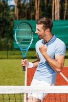 lui conosce lui è il migliore. contento giovane uomo nel polo camicia Tenere tennis racchetta e Esprimere a gesti mentre in piedi su tennis Tribunale foto