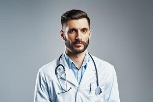 bello giovane uomo nel bianca laboratorio cappotto guardare a telecamera mentre in piedi contro grigio sfondo foto