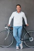 io amore mio bicicletta fiducioso maturo uomo pendente a il suo bicicletta e sorridente mentre in piedi contro grigio sfondo foto