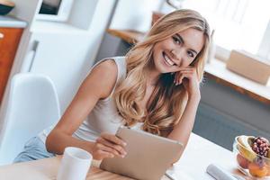 bellezza con digitale tavoletta. bellissimo allegro giovane donna Tenere sua touchpad e guardare a telecamera con Sorridi mentre seduta a il cucina a casa foto