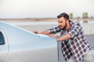 auto guasto. concentrato giovane uomo spingendo il suo auto lungo il strada foto