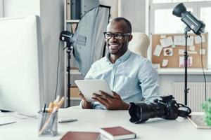 contento giovane africano uomo utilizzando digitale tavoletta e sorridente mentre Lavorando nel il moderno ufficio foto