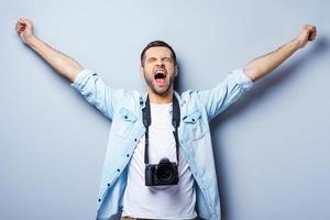 riuscito fotografo. contento giovane uomo con digitale telecamera conservazione braccia sollevato e occhi chiuso mentre in piedi contro grigio sfondo foto