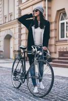 grande tempo metereologico per un' poco giro. bellissimo giovane donna nel occhiali da sole rotolamento sua bicicletta e guardare lontano con Sorridi mentre a piedi all'aperto foto