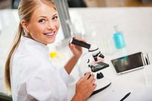 bello giorno nel il laboratorio. superiore Visualizza di sorridente giovane femmina scienziato utilizzando microscopio e guardare a telecamera mentre Lavorando nel il laboratorio foto