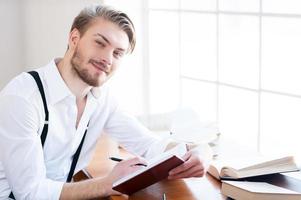 ispirato autore. bello giovane uomo nel camicia e giarrettiere scrittura qualcosa nel Nota tampone e sorridente a telecamera mentre seduta a il suo Lavorando posto foto