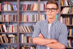 inteligente e fiducioso alunno. bello giovane uomo Tenere libri e sorridente mentre in piedi nel biblioteca foto