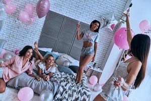 bicchiere di Champagne quattro attraente giovane sorridente donne nel pigiama bonding insieme mentre avendo un' sonno festa nel il Camera da letto con palloncini tutti al di sopra di il posto foto