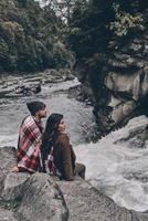 sensazione confortevole insieme. bellissimo giovane coppia coperto con coperta guardare lontano mentre seduta su il rocce vicino il fiume foto