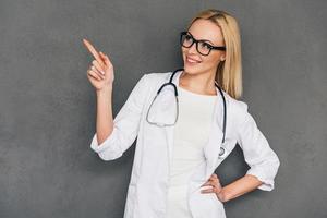 famiglia medico raccomanda voi bellissimo giovane femmina medico puntamento su copia spazio e guardare lontano con Sorridi mentre in piedi contro grigio sfondo foto