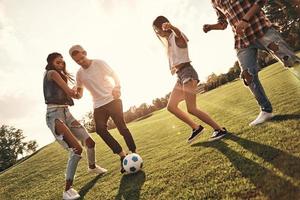 spensierato soleggiato giorno. gruppo di giovane sorridente persone nel casuale indossare sorridente mentre giocando calcio all'aperto foto