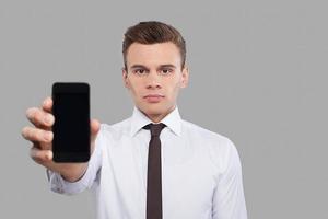 uomo con mobile. bello giovane uomo nel camicia e cravatta mostrando mobile Telefono per telecamera mentre in piedi contro grigio sfondo foto