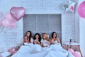 appena rilassante. quattro attraente giovane donne nel pigiama potabile cocktail mentre dire bugie nel il letto con palloncini tutti al di sopra di il camera foto