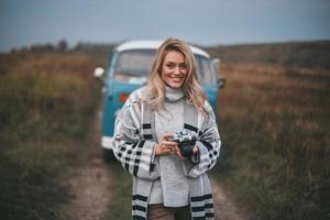 pronto per prendere bene colpi. attraente giovane donna Tenere digitale telecamera e sorridente mentre in piedi all'aperto con il blu retrò stile mini furgone nel il sfondo foto