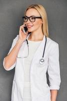 bene parlare con sua paziente bellissimo giovane femmina medico parlando su mobile Telefono con Sorridi mentre in piedi contro grigio sfondo foto