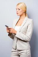grave Messaggio conversazione. bellissimo giovane donna d'affari Tenere mobile Telefono e guardare a esso mentre in piedi contro grigio sfondo foto