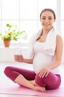 rilassante dopo esercizio. bellissimo incinta donna seduta nel loto posizione e Tenere un' bicchiere con acqua foto